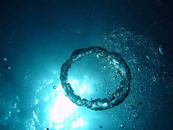 Air ring blown in dark blue water with sun above surface level shining through water.