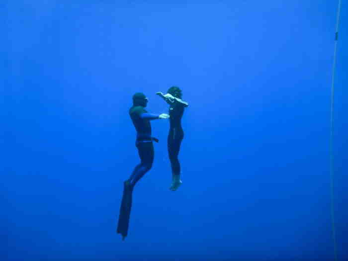 Freediving instructor coaching another freediver underwater in light blue water.